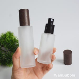May include: Two cylindrical frosted glass bottles with a smooth finish. One bottle features a dark brown wood-grain cap, while the other has a black spray pump nozzle. A separate matching wood-grain cap sits nearby. The text WenBubble is visible.