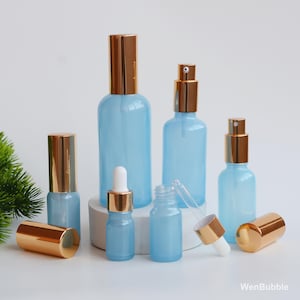 Puede incluir: Colección de botellas de vidrio azul claro con tapas y pulverizadores dorados. Incluye varios tamaños, algunos con cuentagotas. Las botellas están dispuestas sobre una superficie blanca.
