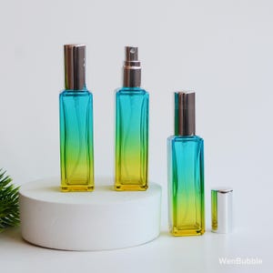 May include: Three rectangular glass perfume bottles with a gradient of turquoise to yellow. The bottles have silver-colored spray tops and caps. One cap is removed and placed next to a bottle. The bottles are on a white surface.