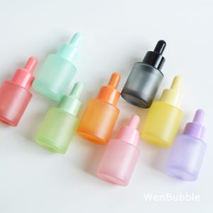 May include: Assortment of frosted glass dropper bottles in various pastel colors, including pink, green, orange, yellow, and purple. Each bottle has a matching colored dropper top, except for one with a black top. The bottles are arranged on a white surface.