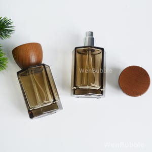 May include: Two rectangular perfume bottles made of translucent brown glass with silver spray nozzles. One bottle has a round wooden cap attached, while the other has a matching wooden cap placed separately. The text WenBubble is visible.