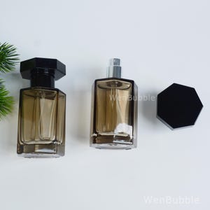 Puede incluir: Dos frascos de perfume de vidrio vacíos con un tono marrón ahumado y formas hexagonales. Un frasco tiene una tapa hexagonal negra puesta, mientras que el otro se muestra sin tapa, revelando un atomizador plateado. El texto WenBubble es visible.