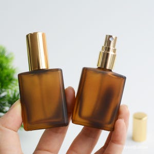May include: Two amber glass perfume bottles with gold-colored spray tops. One bottle has the cap on, and the other has the spray nozzle exposed. The bottles are rectangular with rounded corners. A gold cap sits to the side.
