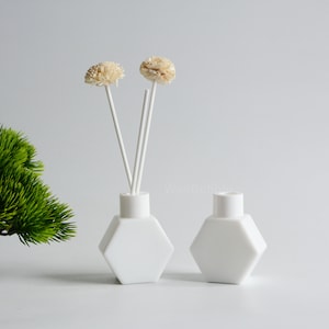 Könnte beinhalten: Zwei weiße Keramik-Diffuserflaschen mit sechseckigen Basen. Die Flasche links hat weiße Diffusorstäbchen und getrocknete weiße Blüten.