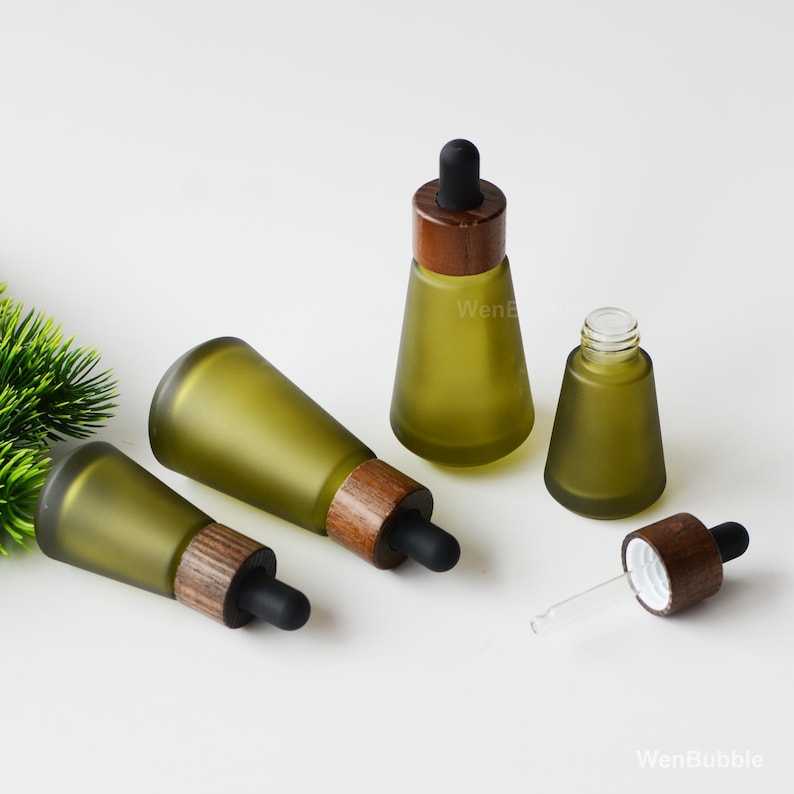 May include: Olive green glass dropper bottles with wooden caps and black rubber droppers. The bottles are in various positions, some upright and some lying down. One bottle is open, showing the glass dropper. The bottles are on a white surface.