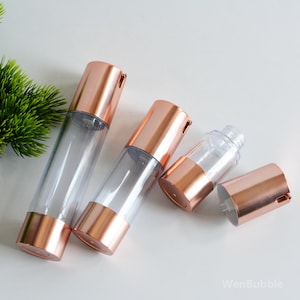 May include: Four clear plastic bottles with rose gold coloured tops and pumps. The bottles are different sizes and are arranged on a white surface.