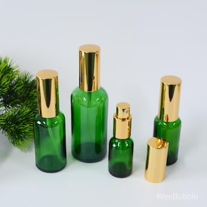 Op de afbeelding: Set van vijf groene glazen flessen met gouden spuitkoppen. De flessen zijn van verschillende grootte en zijn in een rij op een wit oppervlak gerangschikt.