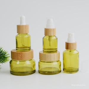 Könnte beinhalten: Eine Sammlung grüner Glasflaschen mit Bambusdeckeln und weißen Applikatoren. Das Set umfasst eine Tropfflasche, eine Sprühflasche und Gläser in verschiedenen Größen. Die Flaschen sind auf einer weißen Oberfläche angeordnet.