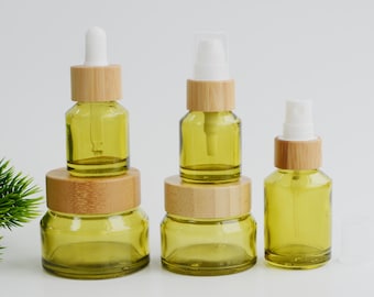 May include: A collection of green glass bottles with bamboo lids and white applicators. The set includes a dropper bottle, a spray bottle, and jars of varying sizes. The bottles are arranged on a white surface.