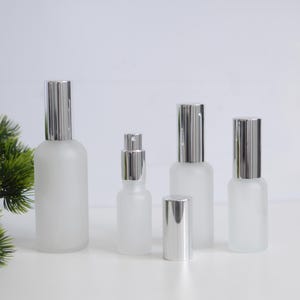 May include: A collection of frosted glass spray bottles with silver caps. The bottles are of varying sizes, with a small one in the centre and larger ones on either side. The caps are cylindrical and shiny, and the bottles are on a white surface.