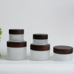 Peut inclure: Plusieurs pots en verre dépoli avec des couvercles en bois brun foncé. Les pots sont de différentes tailles, empilés et disposés sur une surface blanche. Les couvercles ont un aspect de grain de bois texturé. Les pots sont vides.