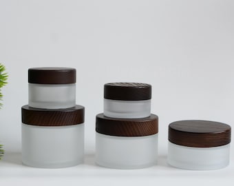 Op de afbeelding: Een verzameling glazen potten met een matte afwerking en donkerbruine houten deksels. De potten zijn in verschillende maten, gestapeld en gerangschikt op een wit oppervlak. De deksels hebben een textuur van houtnerf. De potten zijn leeg.