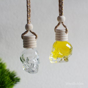 Può includere: Due profumatori per auto a forma di teschio di vetro appesi a una corda. Uno è pieno di liquido giallo e l'altro è vuoto.