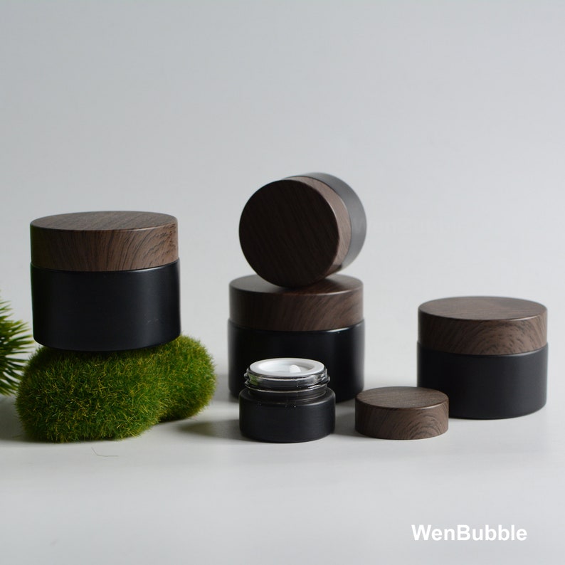May include: Set of five black glass jars with wooden lids. The jars are different sizes and are arranged in a pyramid shape. The jars are perfect for storing cosmetics, lotions, or other small items.