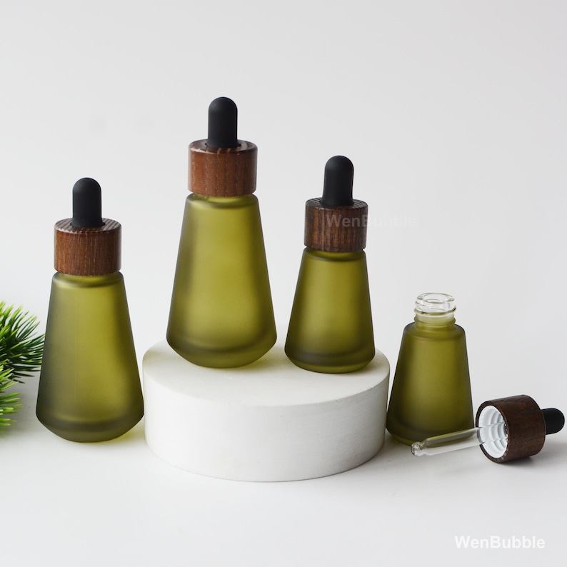 May include: Four olive-green glass dropper bottles with wooden caps and black droppers. One bottle is open, revealing the glass dropper. The bottles are arranged on a white surface, with a small plant in the background.