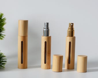 May include: Three bamboo travel spray bottles with different colored atomizers. The bottles are clear glass with a bamboo exterior. The atomizers are silver, gold, and gold. The bottles are all closed and sitting on a white surface.