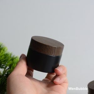 May include: A black glass jar with a wooden lid. The jar is round and has a smooth surface. The lid is made of wood and has a natural grain pattern.