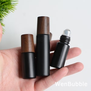 May include: Three black essential oil roller bottles with wooden caps are held in a hand. The bottles vary in size, with the largest in the center. The rollerball is visible on the smallest bottle. The brand name "WenBubble" is visible in the bottom right corner.