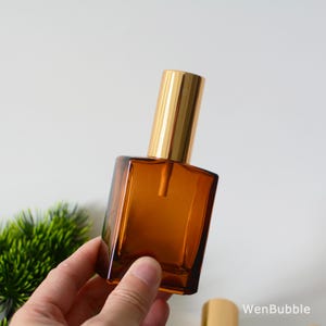 May include: A brown glass bottle with a gold spray top. The bottle is square shaped and has a frosted finish.
