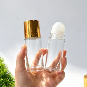 May include: Two clear glass roll-on bottles. One bottle features a shiny gold-colored cap, while the other is shown without a cap, revealing a white plastic roller ball mechanism at the top. Both containers have a cylindrical shape and clear glass.