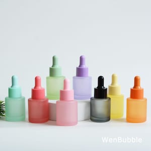 May include: A collection of colorful frosted glass dropper bottles. The bottles are in various pastel shades including pink, green, purple, and yellow. Each bottle has a matching colored dropper top. The bottles are arranged on a white surface.