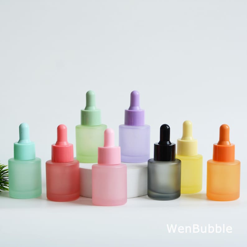 May include: A collection of colorful frosted glass dropper bottles. The bottles are in various pastel shades including pink, green, purple, and yellow. Each bottle has a matching colored dropper top. The bottles are arranged on a white surface.