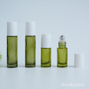 May include: Four olive green glass roller bottles with white caps, arranged in a row. Two tall bottles are on the left, one shorter bottle is in the center, and one bottle with its white cap removed to reveal a metal roller ball is on the right.