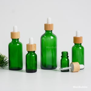 Op de afbeelding: Een verzameling van vier groene glazen druppelflesjes met bamboe kragen en witte rubberen dopjes. Drie flesjes staan rechtop met de pipetten erin, terwijl een kleiner flesje open naast de losse glazen pipet en bamboe dop is geplaatst.