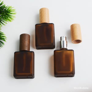 Puede incluir: Tres frascos de perfume de vidrio marrón con tapas de madera se exhiben sobre una superficie blanca. Un frasco tiene un atomizador plateado. Los frascos son rectangulares con esquinas redondeadas. Las tapas de madera son cilíndricas.