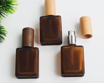 May include: Three brown glass perfume bottles with wooden caps are displayed on a white surface. One bottle has a silver atomizer. The bottles are rectangular with rounded corners. The wooden caps are cylindrical.