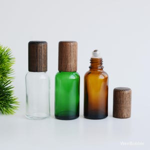 May include: Three glass roller bottles with wooden caps. One is clear, one is green, and the third is amber. The amber bottle has a silver rollerball. The wooden caps are cylindrical and have a wood grain pattern.
