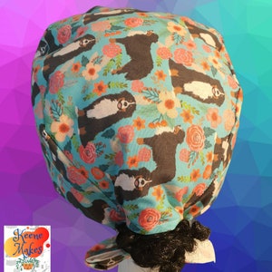 Puede incluir: Un gorro de quirófano azul y blanco con un patrón de perros de montaña de Berna y flores. El gorro está atado en la parte posterior con una cola larga.