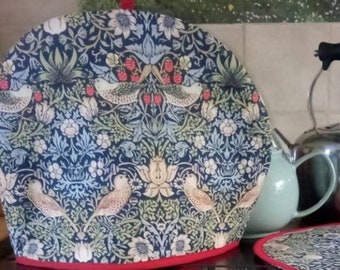 May include: A red and blue patterned tea cozy with a red handle. The pattern features birds, flowers, and leaves. The cozy is sitting on a black surface.