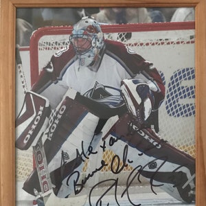 May include: A hockey goalie wearing a maroon and white uniform with a white helmet and a black face mask. The goalie is in a crouched position with a hockey stick in their right hand. The goalie is wearing a black and white goalie pad on their left leg and a black and white goalie pad on their right leg. The goalie is standing on the ice. The goalie's signature is written in black ink on the photo. The signature reads "A.T. to a  Bernie O'Brien".