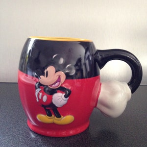May include: A red and black ceramic mug with a Mickey Mouse character design. The mug has a handle shaped like Mickey Mouse's hand.