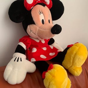 Może przedstawiać: Pluszowa zabawka przedstawiająca Minnie Mouse w czerwonej sukience w białe kropki i z czerwoną kokardką w białe kropki. Ma czarne uszy i czarny nos. Jej oczy są zamknięte i się uśmiecha.
