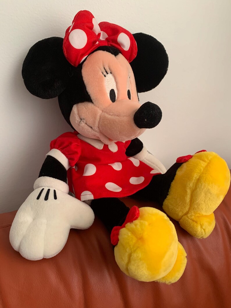 Może przedstawiać: Pluszowa zabawka przedstawiająca Minnie Mouse w czerwonej sukience w białe kropki i z czerwoną kokardką w białe kropki. Ma czarne uszy i czarny nos. Jej oczy są zamknięte i się uśmiecha.
