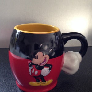 May include: A black and red ceramic mug with a yellow rim and a white handle. The mug features a cartoon image of Mickey Mouse with a red and yellow outfit and a big smile.