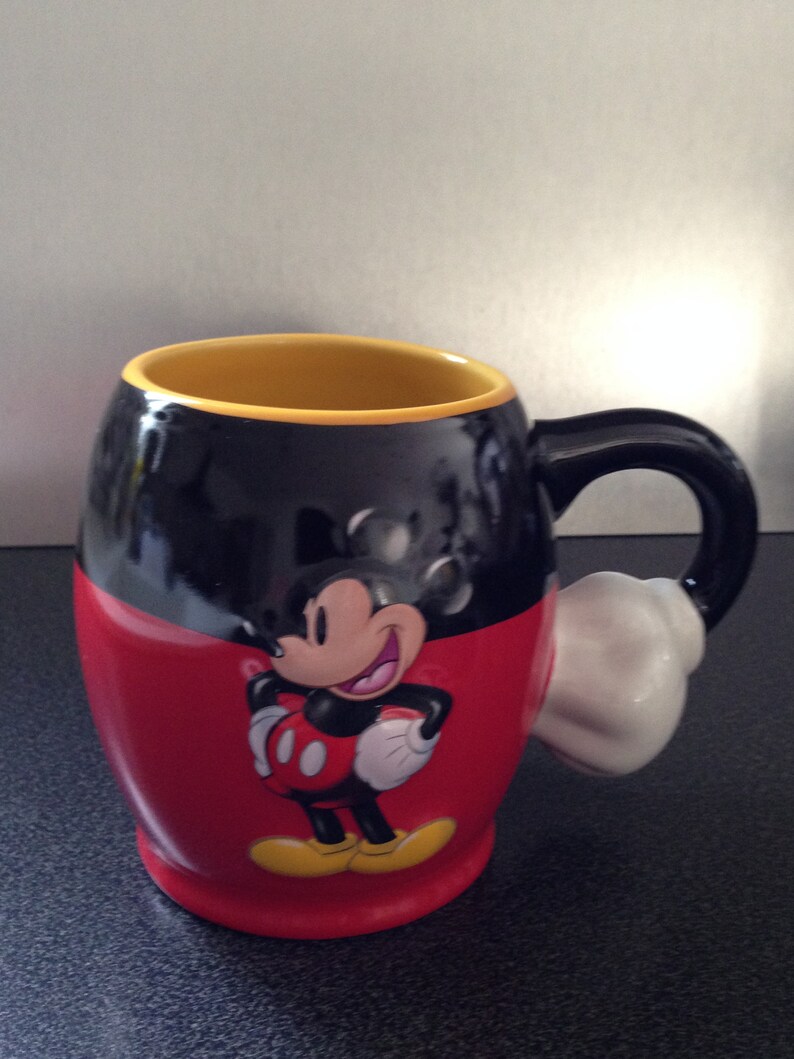 May include: A black and red ceramic mug with a yellow rim and a white handle. The mug features a cartoon image of Mickey Mouse with a red and yellow outfit and a big smile.