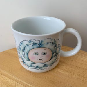May include: White ceramic mug featuring a whimsical illustration of a baby's face nestled within a blue and green cabbage. The mug has a curved handle and a light blue interior, perfect for beverages.