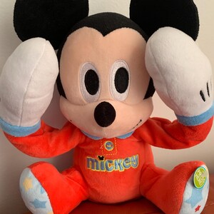 Puede incluir: Un juguete de peluche Mickey Mouse rojo y blanco que lleva un pijama rojo y azul. El juguete tiene un bot&oacute;n en el pecho que dice "Presiona aqu&iacute;".