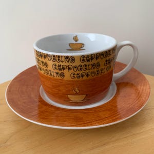 May include: A brown cappuccino cup and saucer set. The cup and saucer have a white interior. The cup is printed with the word "CAPPUCCINO" around the rim. A small coffee cup graphic is on the cup and saucer.