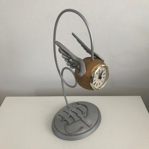 May include: A silver-coloured clock shaped like a Golden Snitch from the Harry Potter series. The clock is on a stand with the Hogwarts crest.