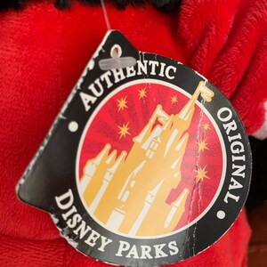 May include: A red plush toy with a black head and a white circle on its ear. The toy has a tag that says "Authentic Original Disney Parks" with a picture of Cinderella's castle.