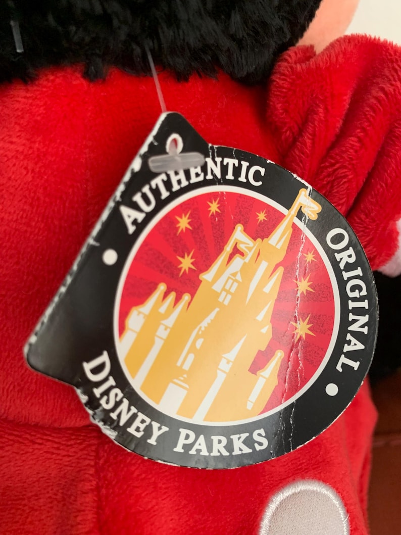 May include: A red plush toy with a black head and a white circle on its ear. The toy has a tag that says "Authentic Original Disney Parks" with a picture of Cinderella's castle.