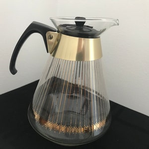 Puede incluir: Una cafetera de vidrio con una banda dorada alrededor de la parte superior y un asa negra. La cafetera tiene un diseño geométrico dorado y negro en la parte inferior.