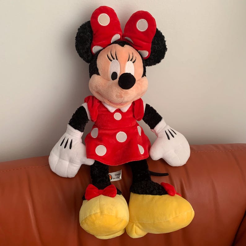 May include: A large plush toy of Minnie Mouse wearing a red dress with white polka dots and a red bow with white polka dots. The toy has black fur and yellow shoes.