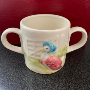 Puede incluir: Taza de cerámica blanca para niños con dos asas. La taza presenta una ilustración colorida de un pato con un gorro azul y un vestido rosa. El pato está de pie frente a una valla verde.