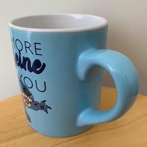 May include: A light blue ceramic mug with a cartoon character on it. The mug has the text "More Caffeine You" on it.