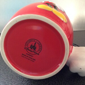 May include: Red ceramic mug with a cartoon image of Mickey Mouse. The mug has a white rim and a black Disney Parks logo on the bottom. The logo reads "Authentic Original Disney Parks &copy;Disney Made in Thailand FAC-014182-15182."